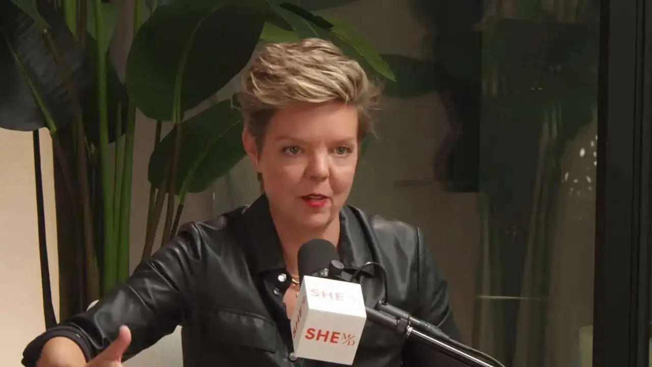Podcast interview guest speaking into a microphone with a clear, well-lit mid-shot and a hand gesture that emphasizes a point.