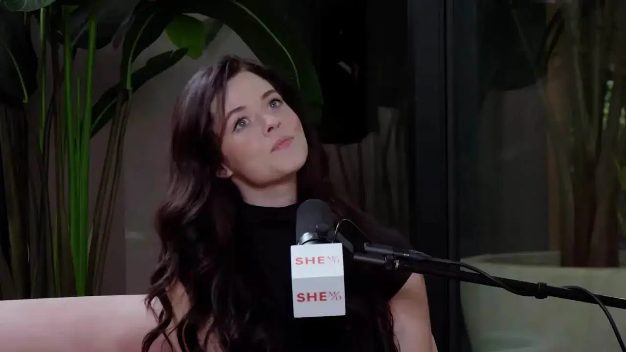 Podcast guest sitting on a sofa speaking into a microphone with a SHE-branded mic flag and plants in the background.