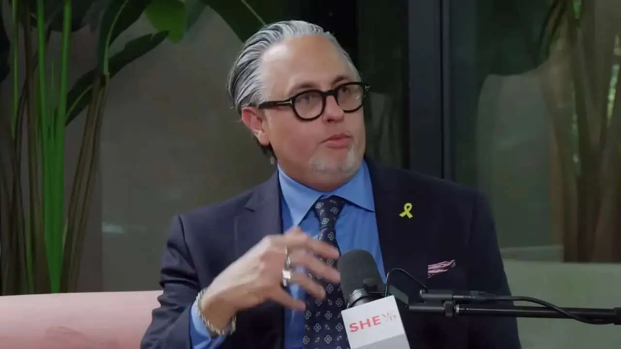 Dr. Thais Aliabadi MD speaking at a press event, wearing a navy suit, glasses, and a yellow ribbon p.