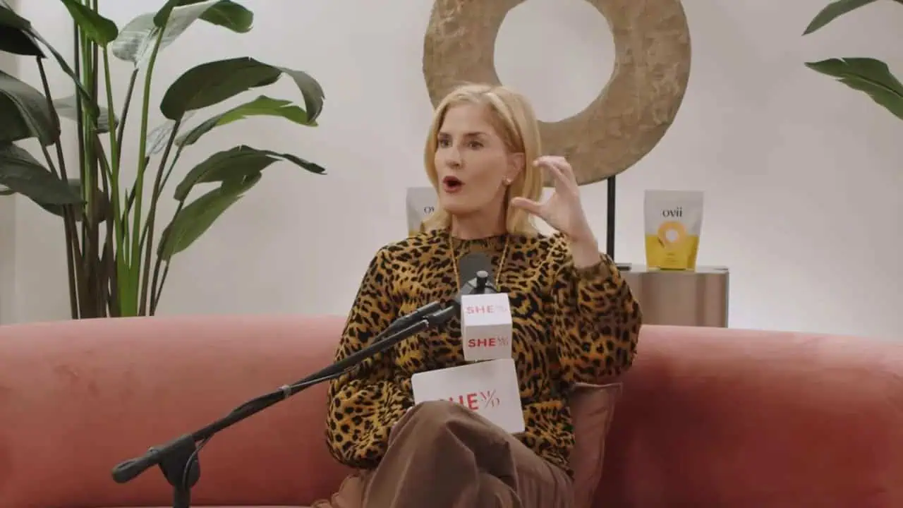 Dr. Thais Aliabadi speaking at an event, seated on a pink sofa with a microphone.