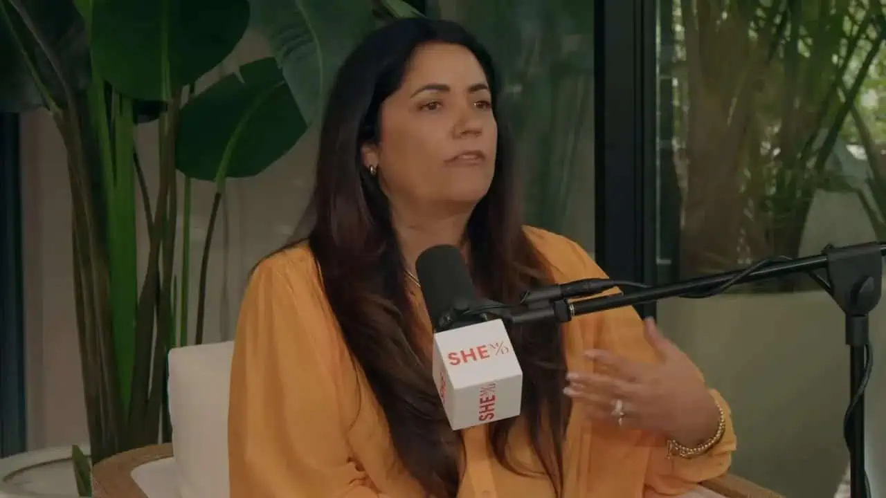 Dr. Thais Aliabadi speaking at a conference or interview, holding a microphone with "SHE" logo, in a.
