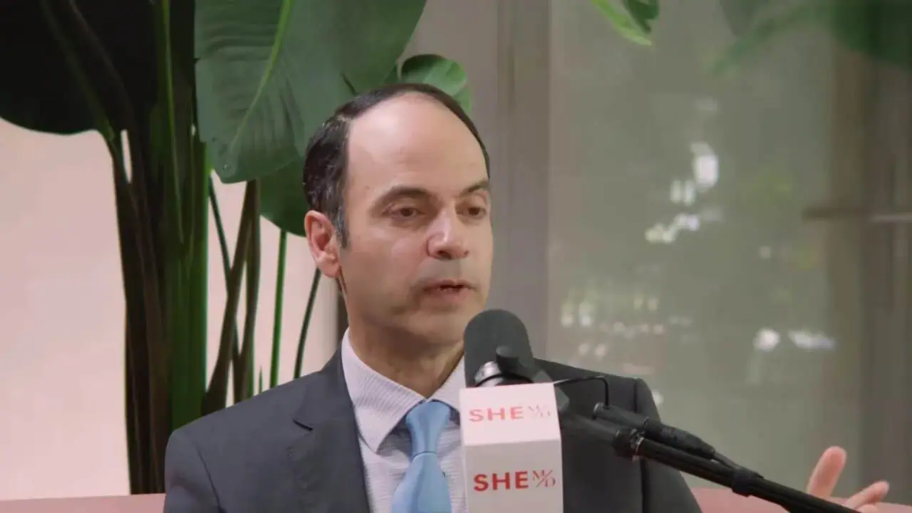 Dr. Thais Aliabadi speaking at an event with a microphone and green plants in background.