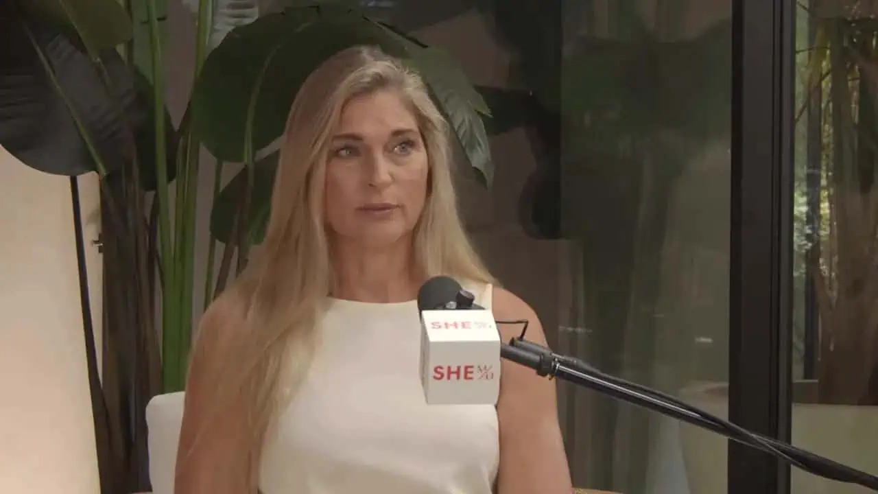 Host seated on a pink couch speaking into a podcast microphone with plants behind her
