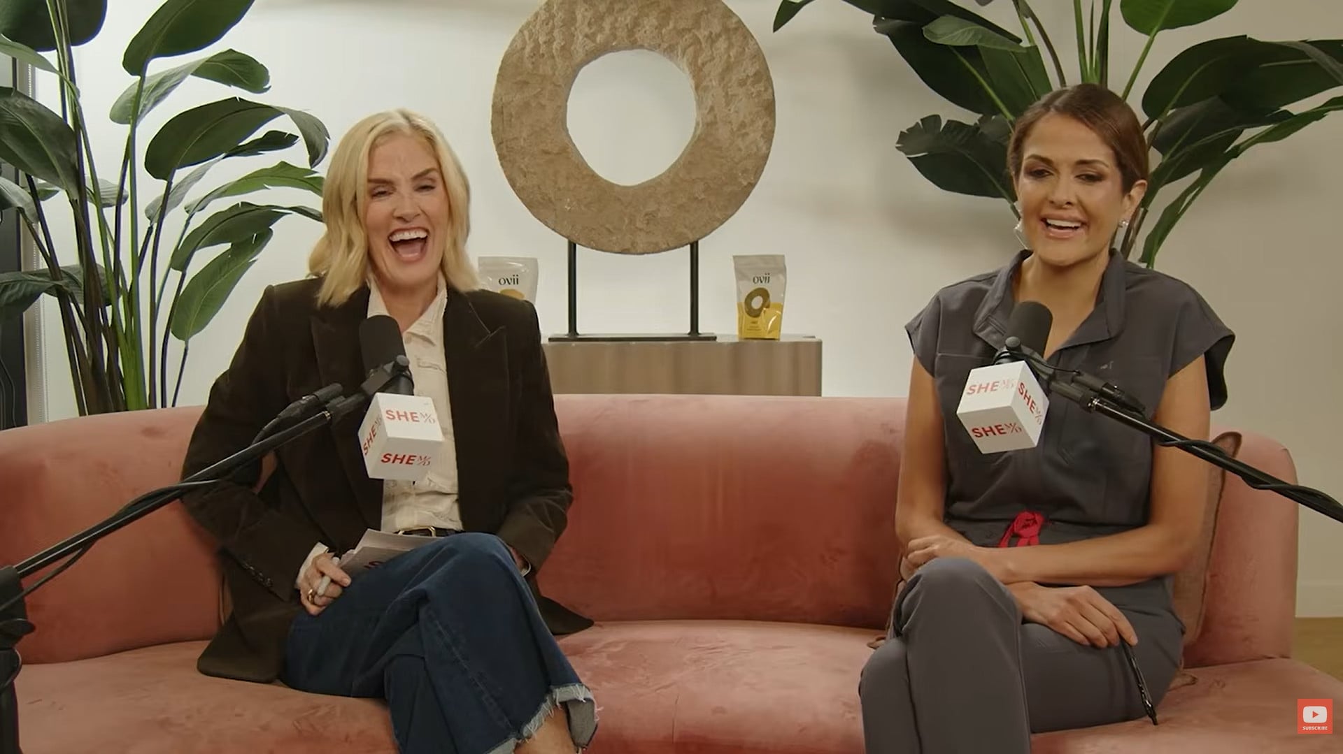 Two women in a podcast interview with microphones and a pink sofa.
