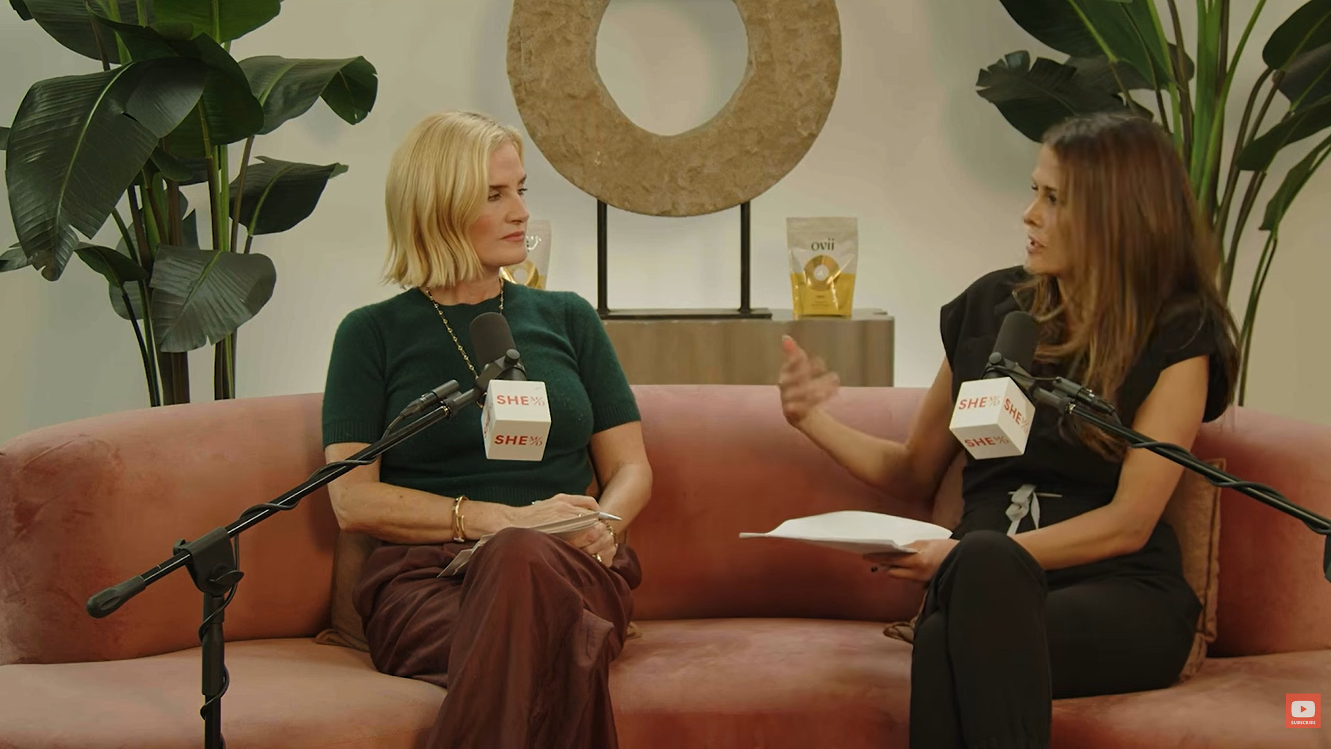 Two women in a podcast interview with microphones and plants in the background.