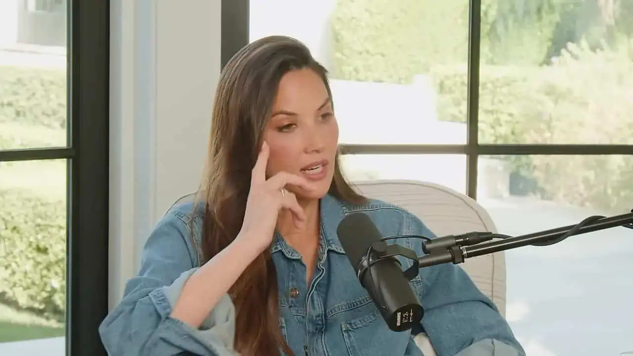 Olivia Munn seated beside a microphone with large windows behind her