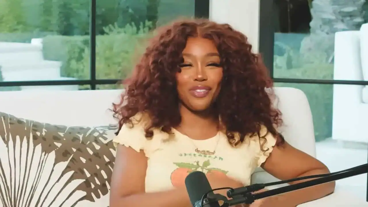 SZA seated in a white chair and gesturing beside a microphone