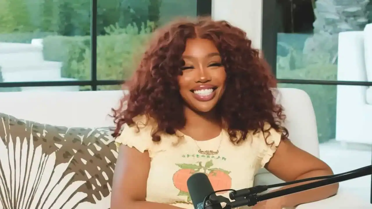 SZA seated in a white chair and gesturing beside a microphone